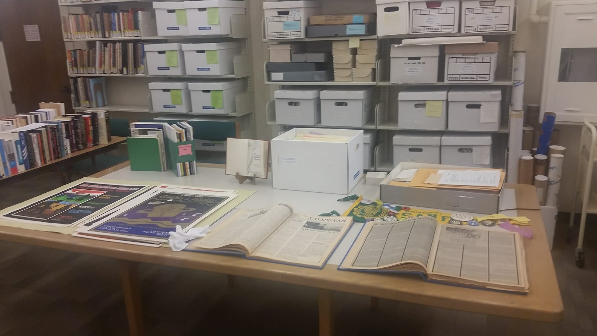 Magazines, books, and posters are spread across a light-colored wood table. There are also some magazine racks and boxes on the table. Behind the table is a shelf of archival boxes and books as well.